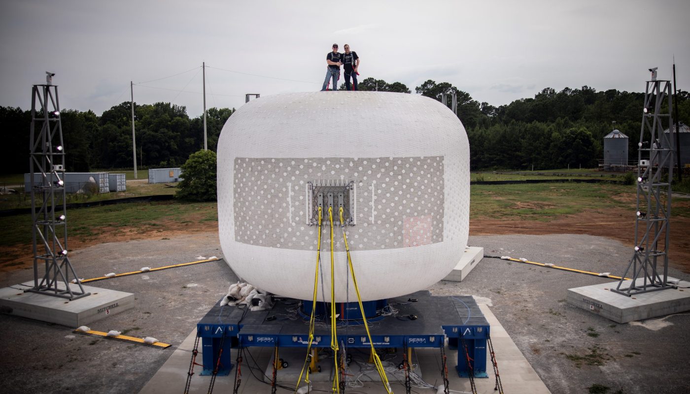 Two people stand on top of a large, white, cylindrical structure secured with yellow straps at a testing site surrounded by greenery and industrial equipment.