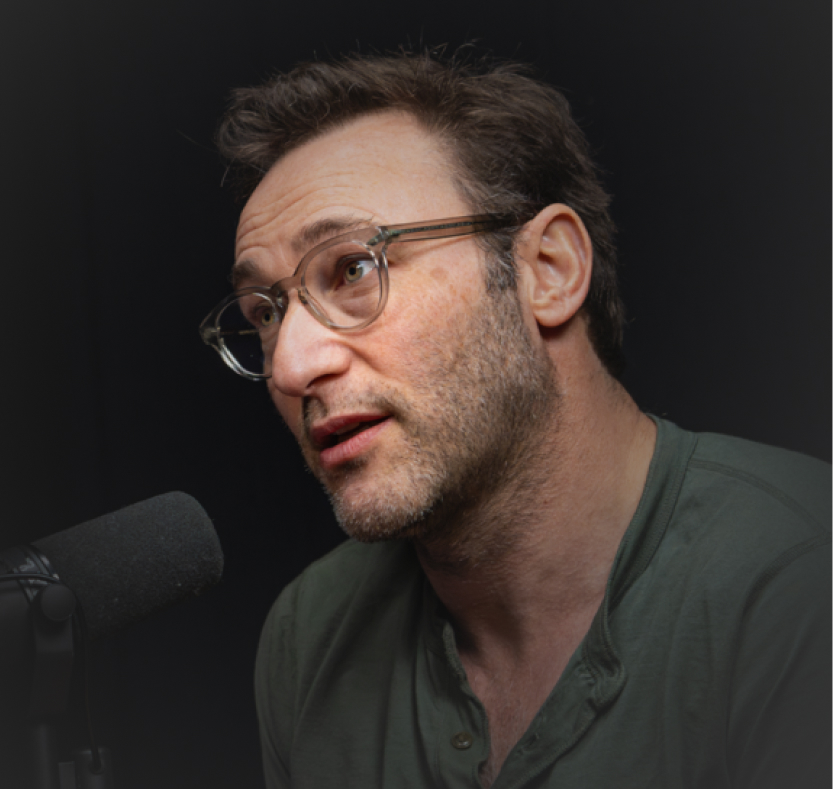 A man with glasses and a beard speaks into a microphone, wearing a green shirt against a dark background.