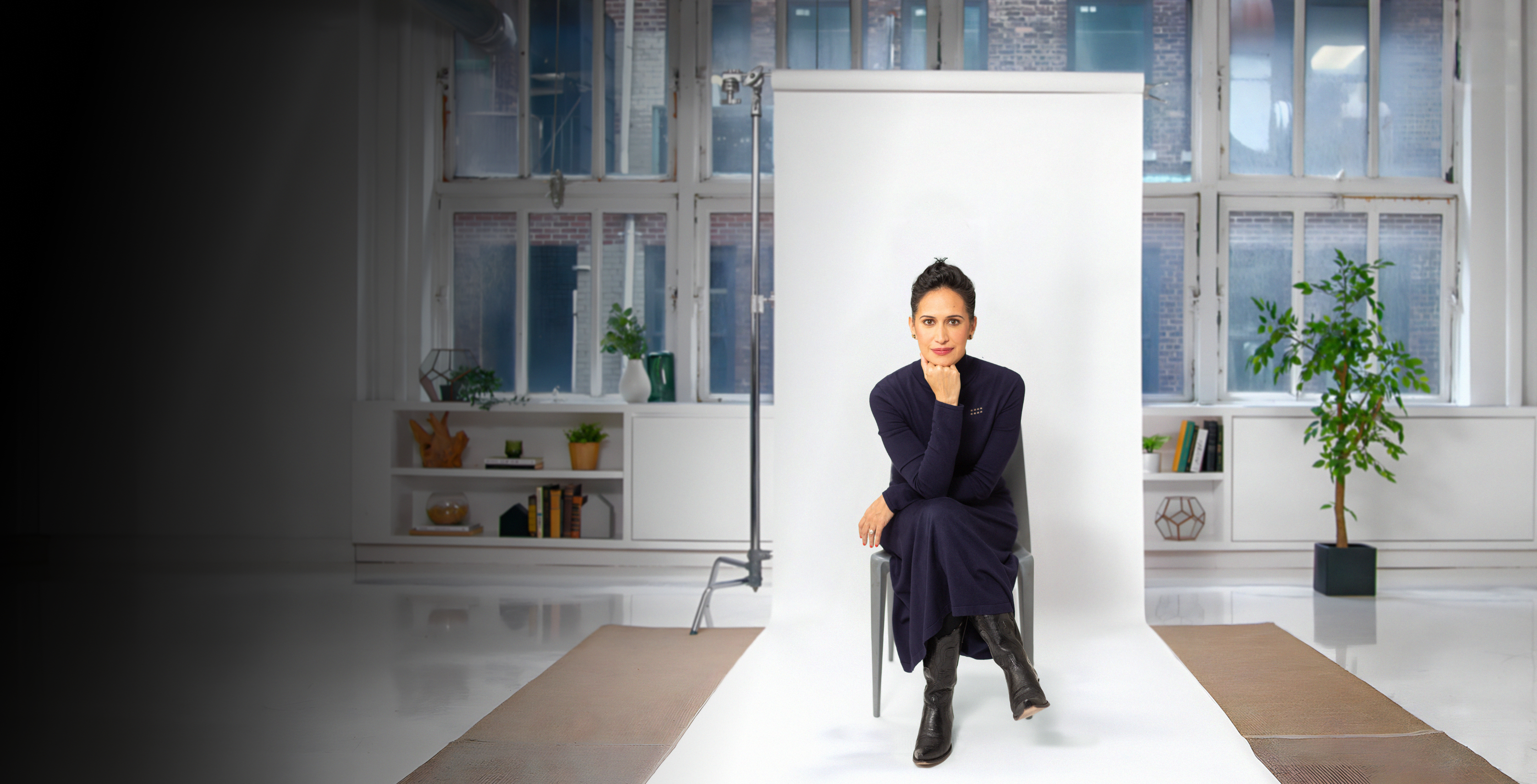 A person in a dark outfit sits on a stool in front of a white backdrop, inside a well-lit studio with large windows and some plants in the background.