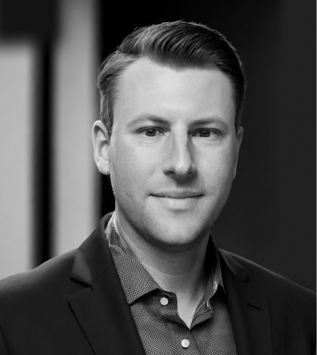 A grayscale portrait of a man with short hair wearing a suit jacket over a patterned shirt, looking directly at the camera.