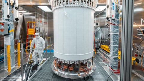 A technician in a cleanroom suit works by a large cylindrical piece of equipment in a high-tech laboratory setting with industrial tools and machinery.