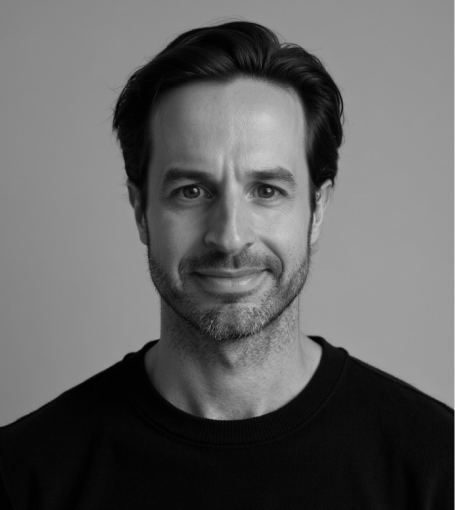 A black-and-white portrait of a smiling man with short, dark hair and a beard, wearing a black sweater.