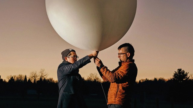 Two individuals are holding and inspecting a large white balloon outdoors, with a silhouette of trees visible in the background at dusk.