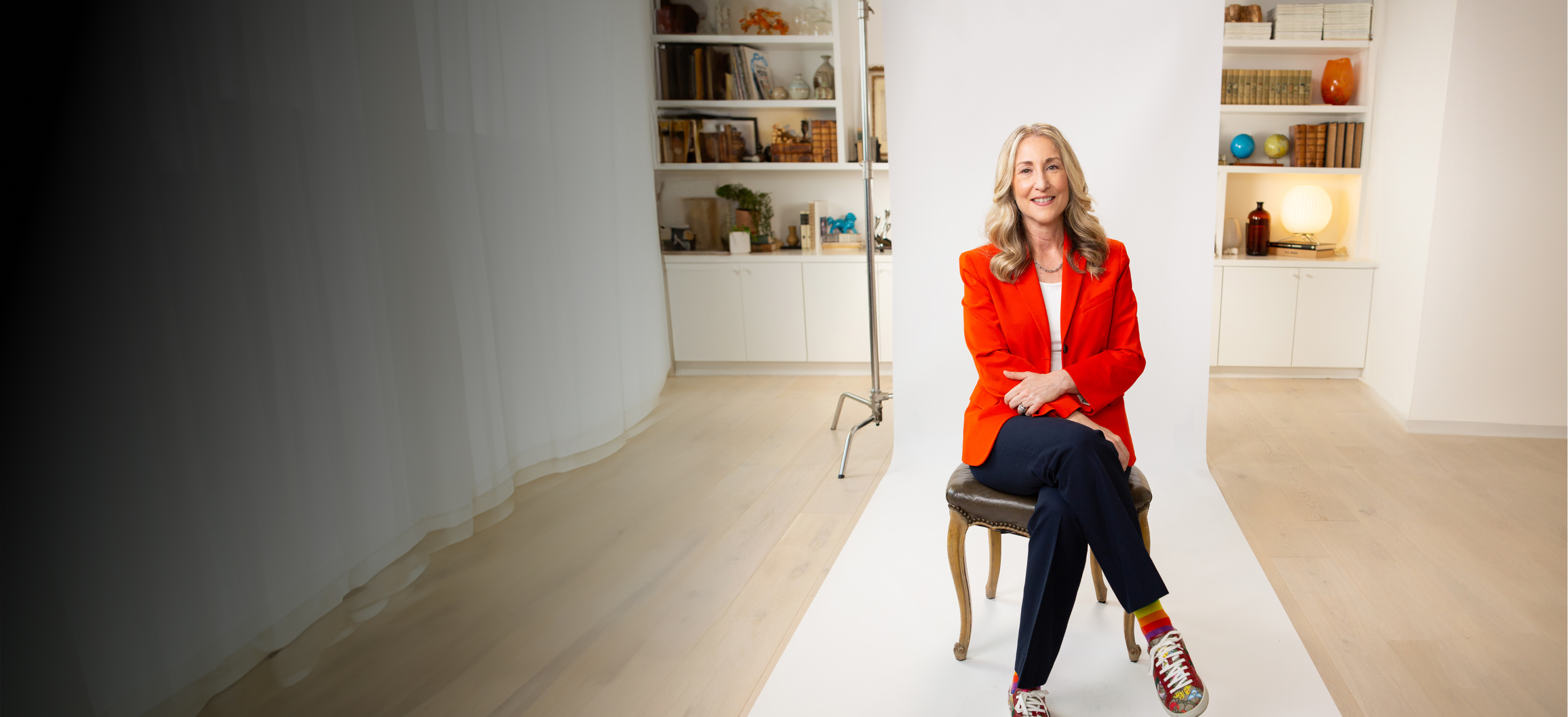 A person with long hair sits on a chair in a photo studio, wearing a red blazer, dark pants, and colorful sneakers. Bookshelves and lighting equipment are visible in the background.