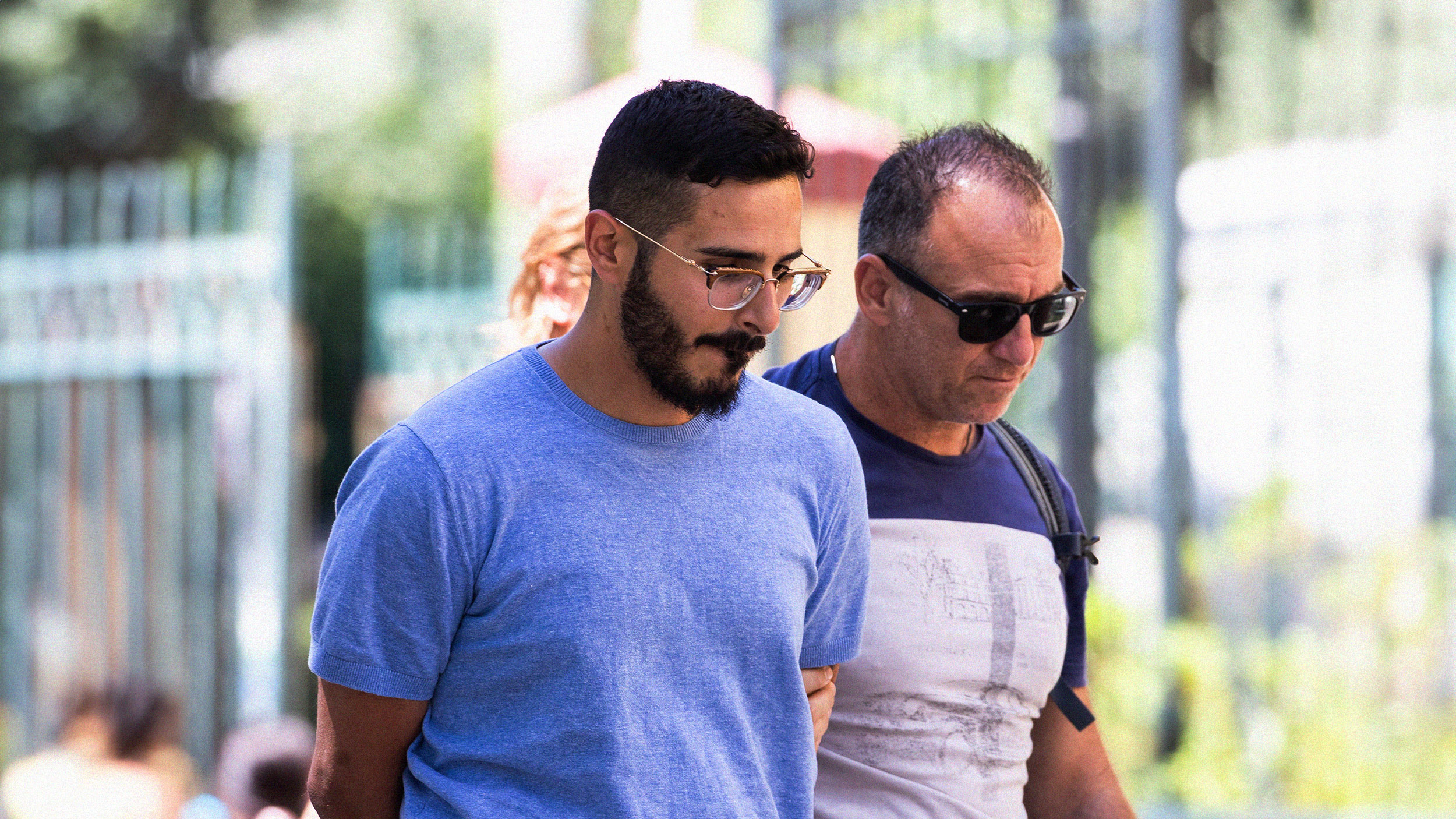 Two men wearing glasses walk side by side outdoors in a sunlit park, with blurred greenery and other people in the background, discussing recent incidents of romance fraud.