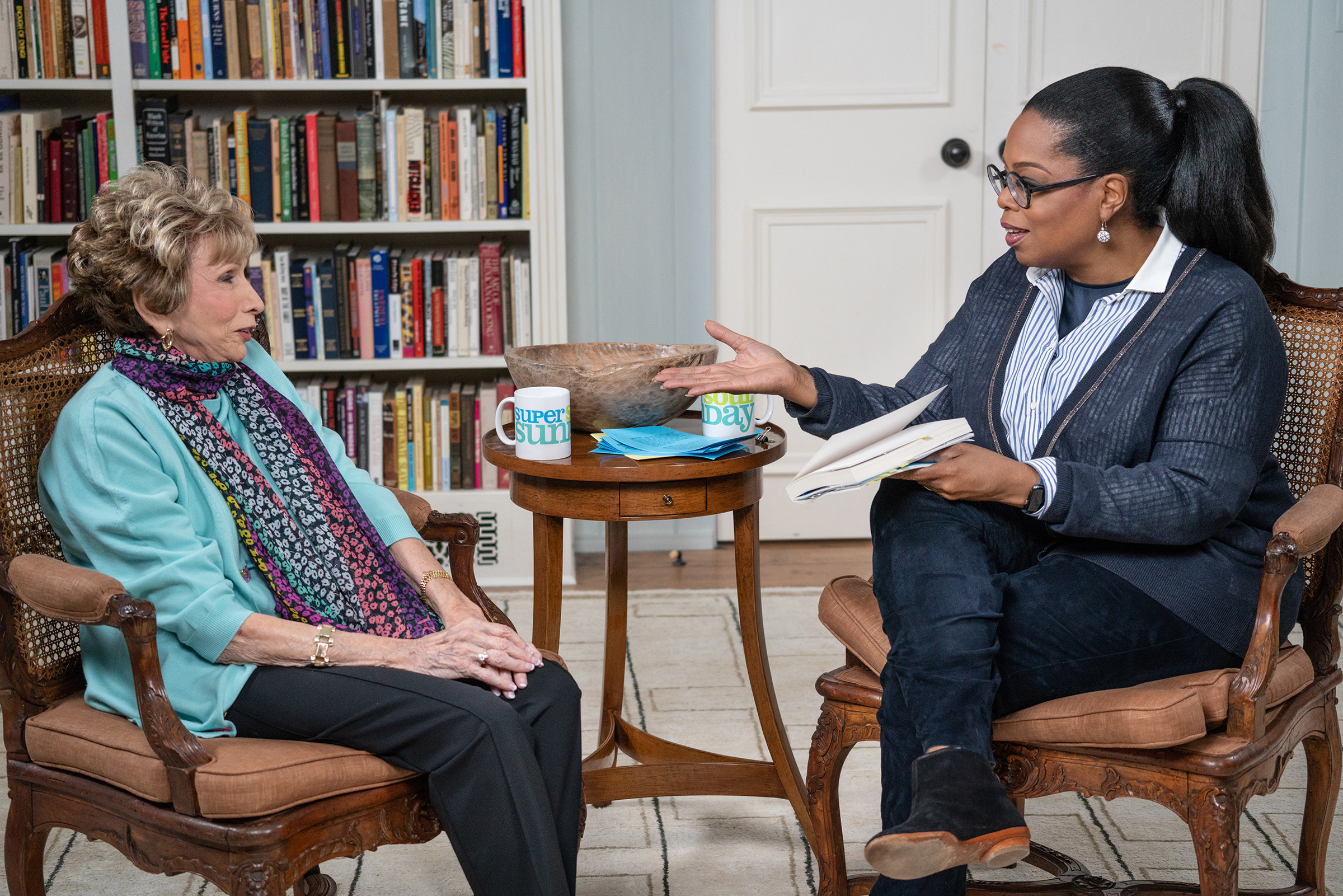 Two people are engaged in conversation in a living room. One is holding a notebook, possibly noting insights inspired by Edith Eva Eger's works. There are two mugs on the table between them, and bookshelves filled with thought-provoking reads are visible in the background.