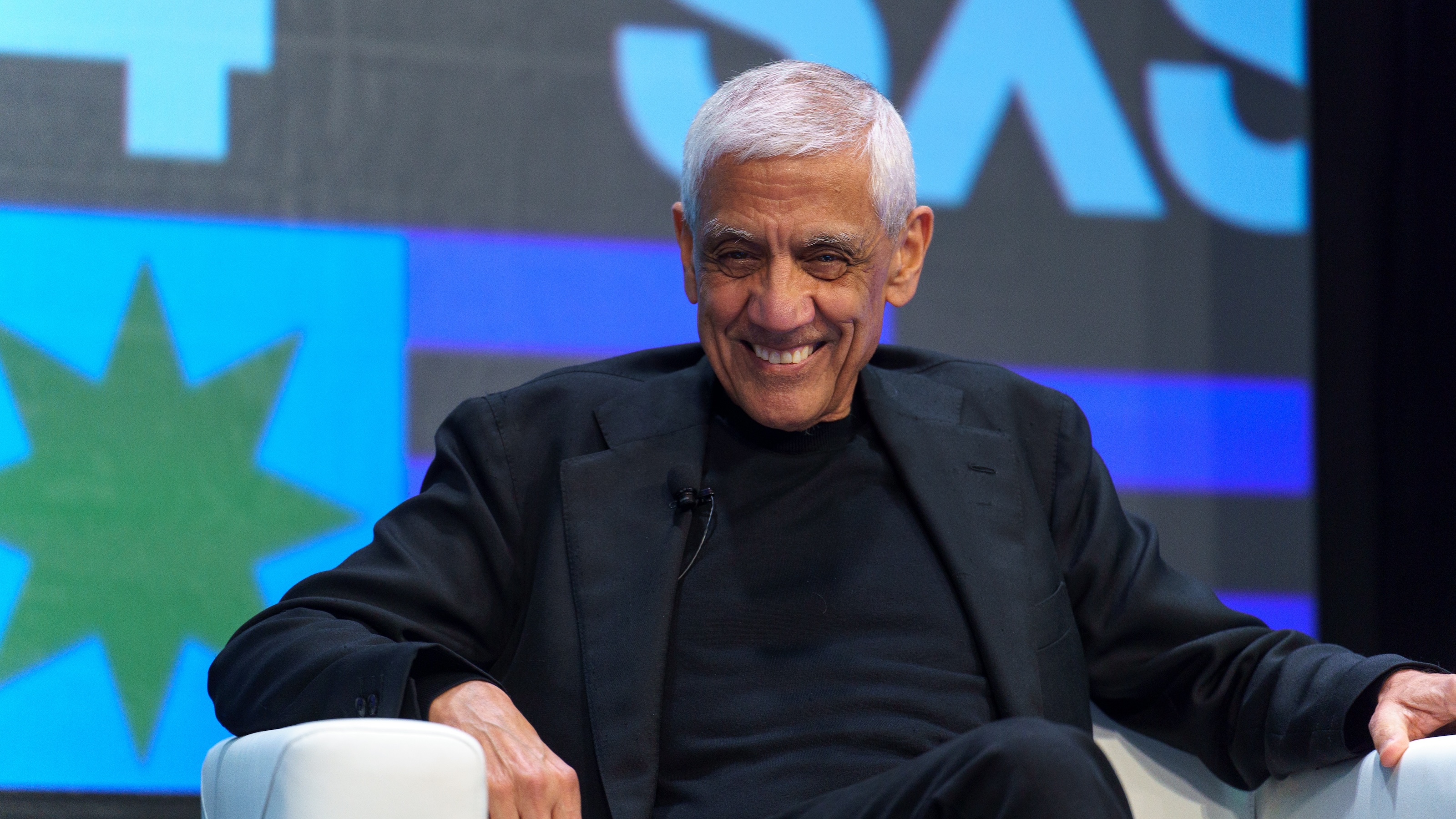 An older man with gray hair is sitting in a white chair, smiling. Dressed in a black outfit, he is framed against a large screen filled with abstract shapes and letters that evoke an AI manifesto.