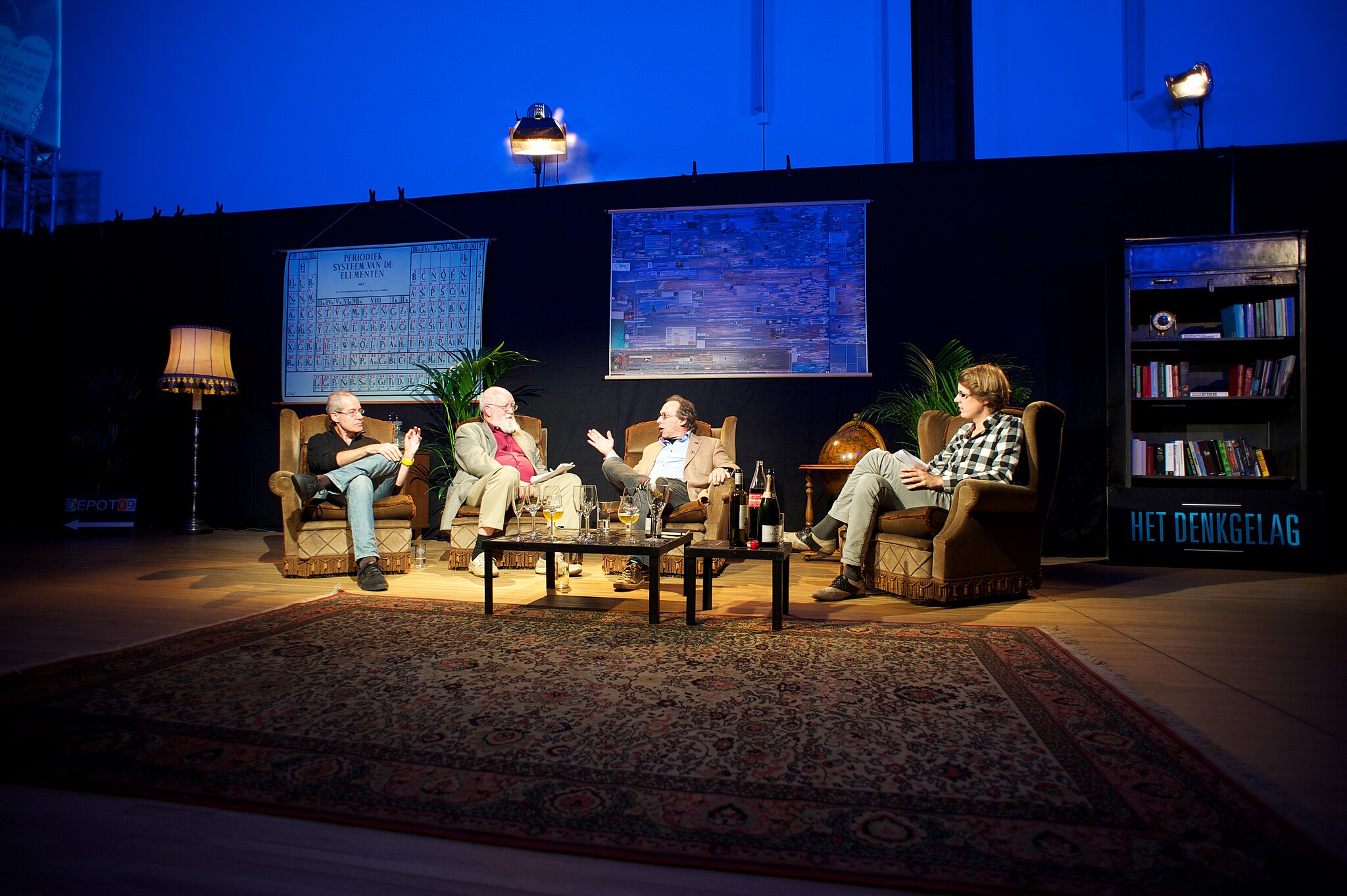 Four people having a panel discussion on stage, seated in armchairs with a rug underfoot, surrounded by plants, bookshelves, and charts on a black backdrop.