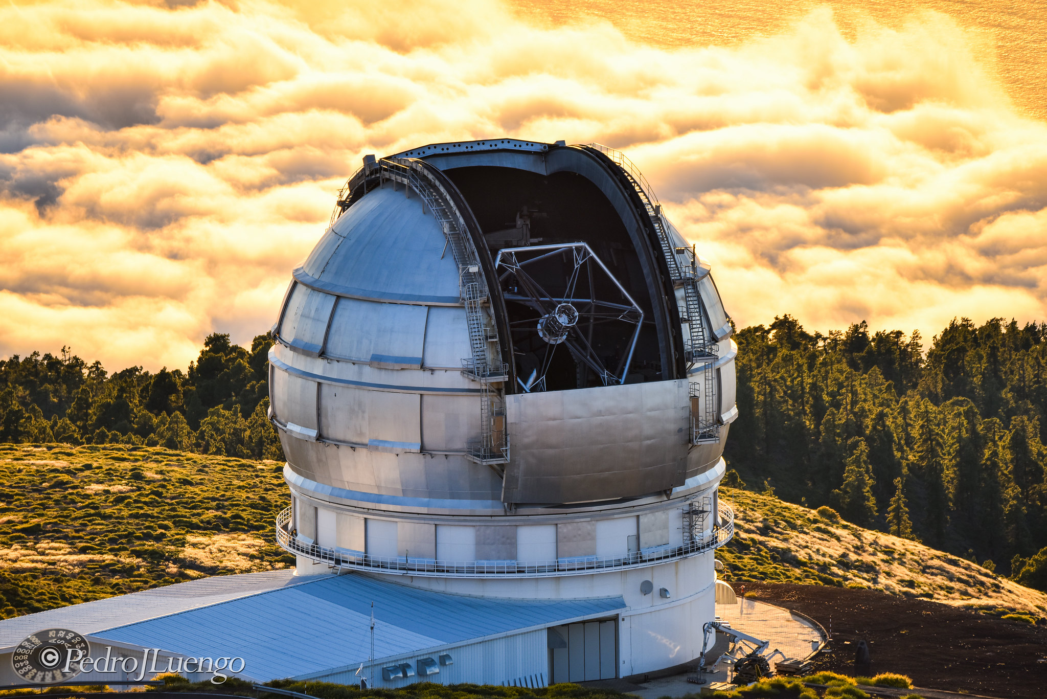 A large observatory telescope with a dome sits on a mountain, hailed as one of the best astronomy locations on Earth, surrounded by trees and clouds during sunset.