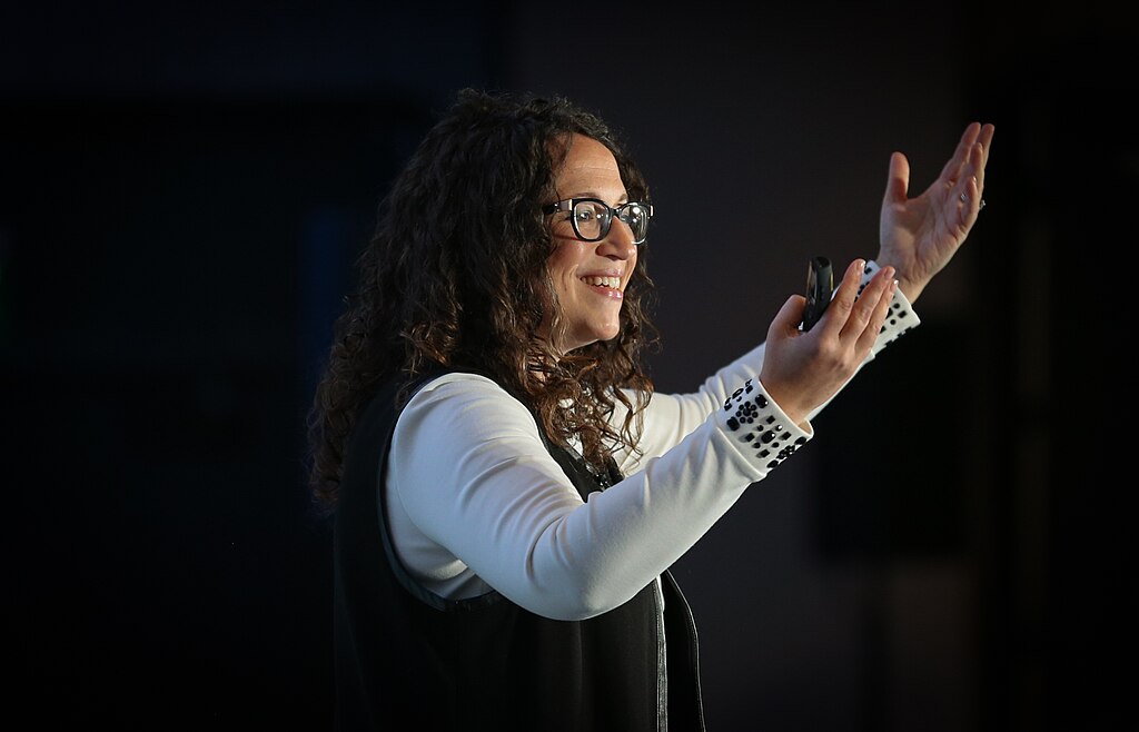 A woman with glasses gestures enthusiastically while speaking, holding a microphone in one hand.