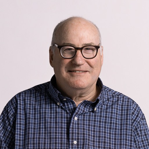 A man with glasses and a checkered shirt smiles in front of a plain white background.