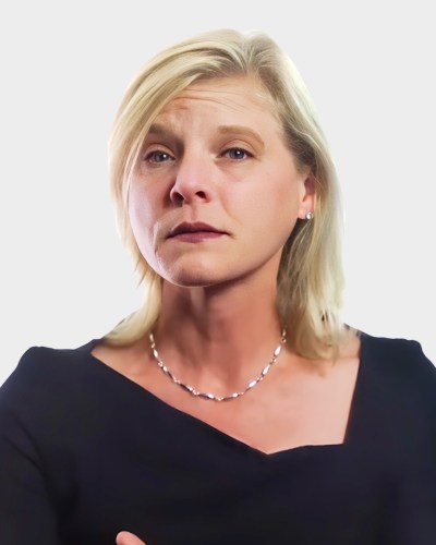 A woman with short blonde hair wearing a black top and a necklace looks slightly upward against a plain background.