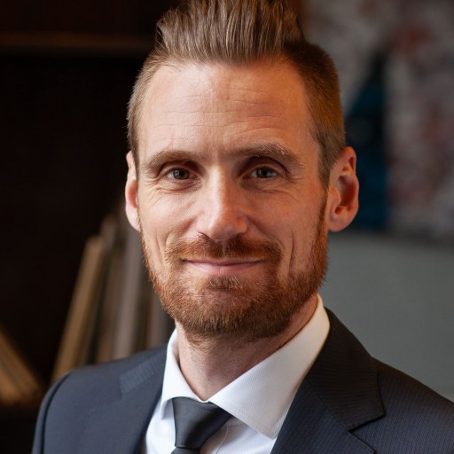 A man with short hair and a beard wears a suit and tie, smiling slightly, in an indoor setting.