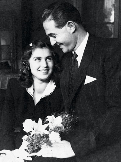 A woman holding flowers looks up at a man in a suit who is smiling at her. They are indoors.