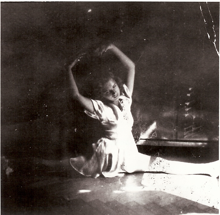 Black and white photo of a woman seated on the floor with arms raised above her head, wearing a dress and smiling. The scene is softly lit, casting shadows around her.