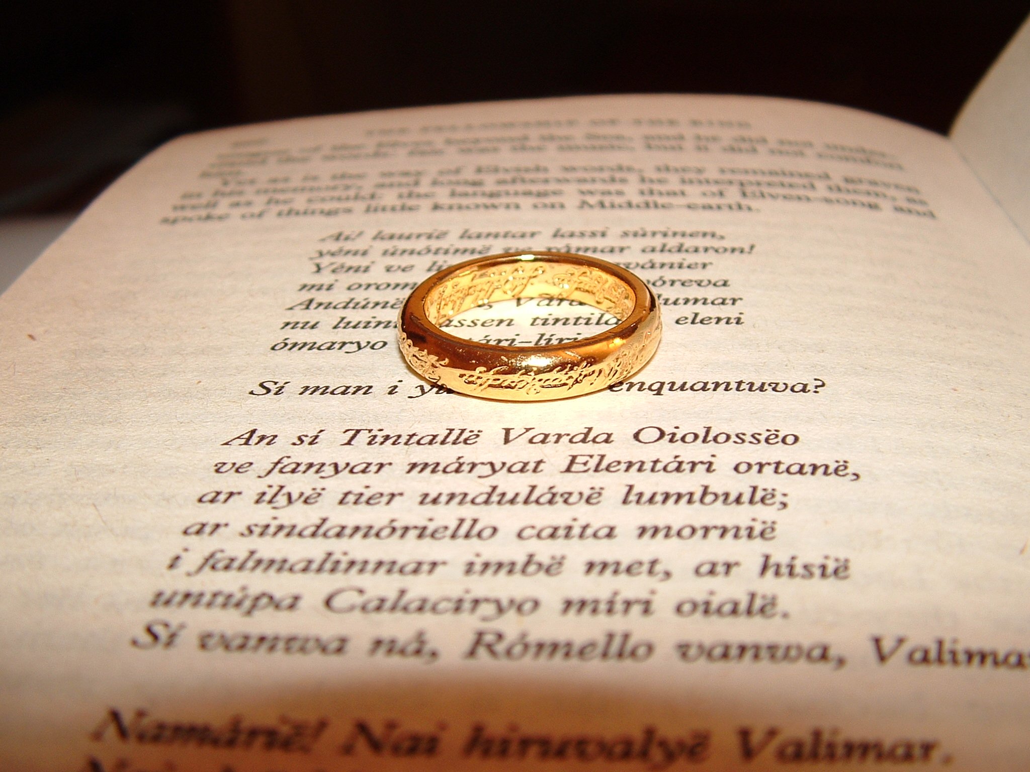 A golden ring sits atop an open book with text written in an elven script, creating a bright reflection on the page.