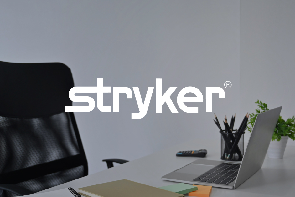 Office desk with a laptop, plant, pens, notebooks, and a TV remote, featuring the Stryker logo in the center.