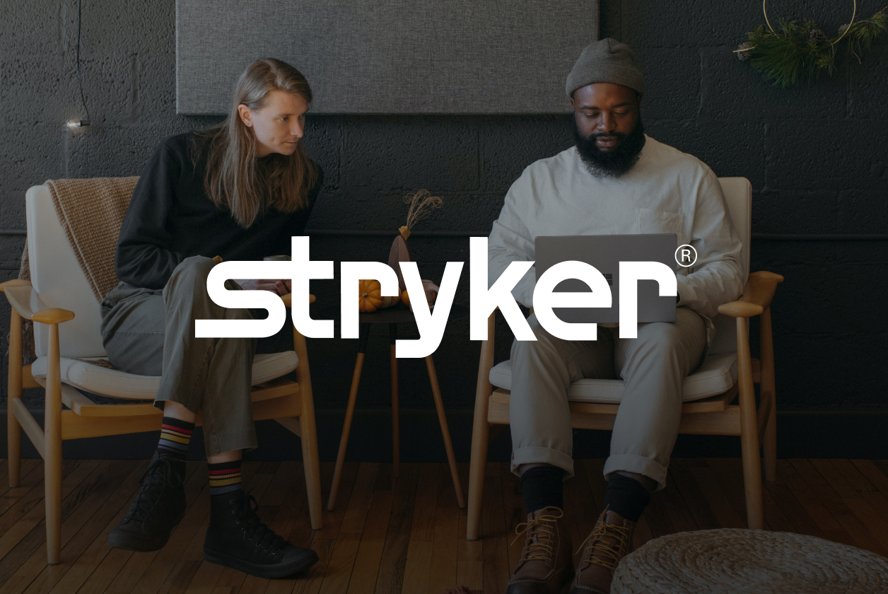 Two people sitting in chairs, one with a laptop, in a cozy room with wooden floor and dark walls. The word "Stryker" is overlaid on the image.