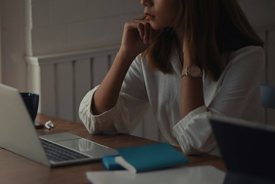 Woman in a white shirt sitting at a table, looking at a laptop with a notebook and mug nearby.