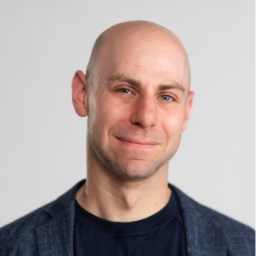 A bald man in a dark blazer and black shirt smiles gently against a plain light background.