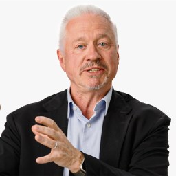 A man with gray hair and a beard, wearing a black jacket and blue shirt, gestures with his hands in front of a plain background. There's a logo in the top right corner.