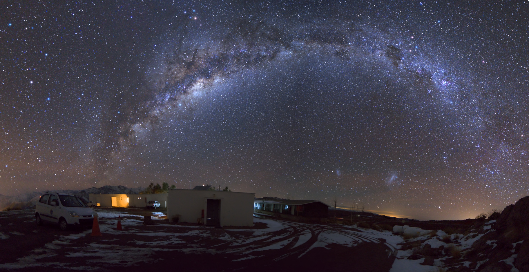 Panoramic view of the Milky Way arching over a small building and vehicles in a snowy landscape at night, showcasing one of the best astronomy locations on Earth.