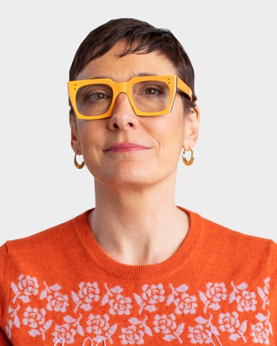 Person with short hair wearing large yellow glasses, a red patterned sweater, and hoop earrings, against a plain background.