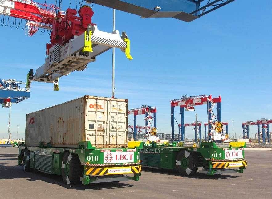 Self-driving trucks transport a shipping container at a port with cranes in the background.