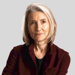 An older person with short grey hair and a maroon jacket looks at the camera against a grey background.