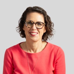 Woman with short curly hair and glasses wearing a coral top, smiling against a light gray background.