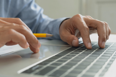 Close-up of hands typing on a laptop keyboard, with one hand holding a pencil.