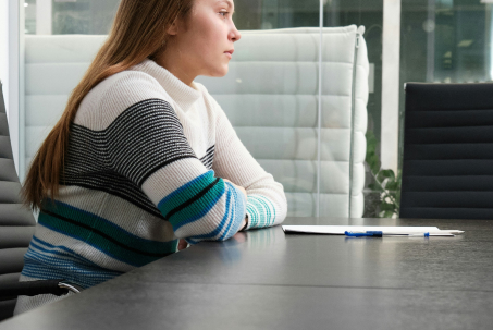 A person with long hair sits at a conference table with arms crossed, looking to the side. A clipboard and pen are on the table.