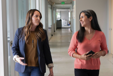 Two women walking and talking in an office hallway, one holding a phone.