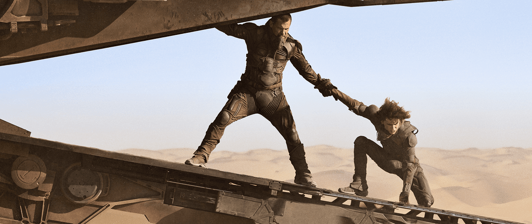 Two people in futuristic attire are on a large vehicle in a desert. One is standing and helping the other climb up. The sky is clear.