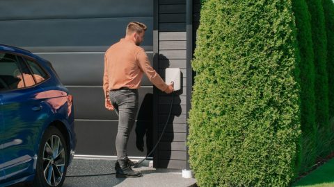 A man connects an electric car to a charging station mounted on a house wall next to a large shrub.