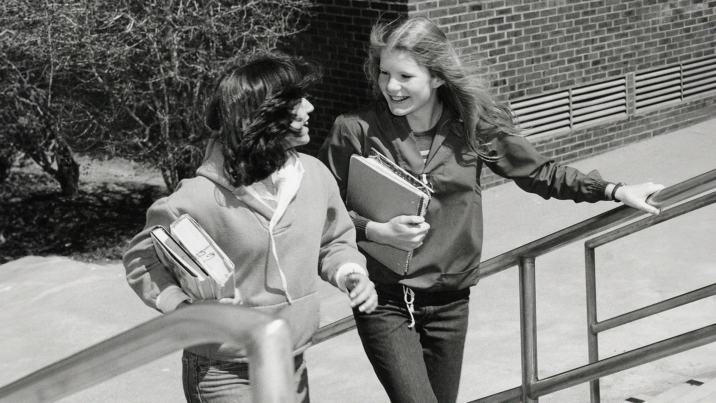 Two friends walking up outdoor stairs, holding books and smiling at each other, their camaraderie evident in every step.