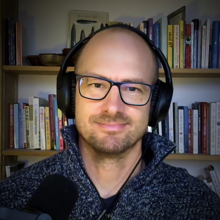 A person wearing glasses and headphones stands in front of a bookshelf with a microphone visible.