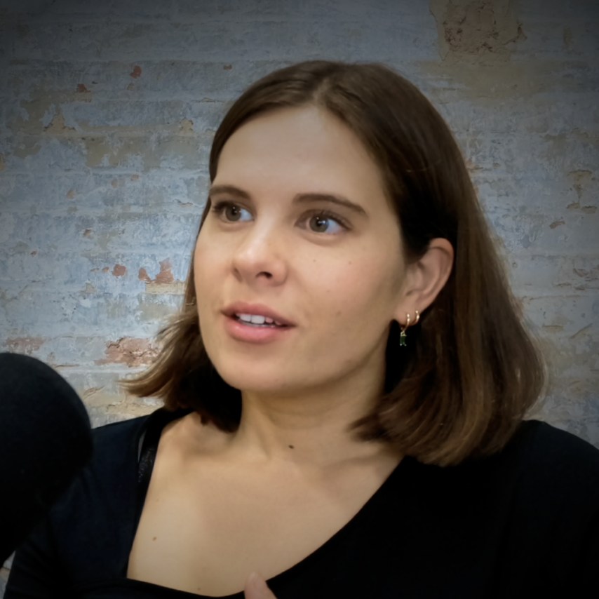 A woman with shoulder-length brown hair is speaking into a microphone. She is wearing a black top and earrings, with a textured wall in the background.