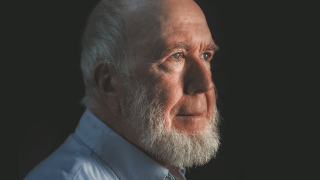 A side profile of an older man with a white beard, wearing a light blue shirt, against a dark background, illuminated by soft lighting from the left, exudes an aura of radical optimism.