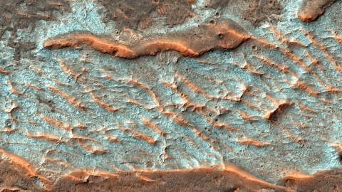 Aerial image of a Martian landscape with rough, textured surface featuring blue and reddish-brown hues marked by undulating ridges and valleys.