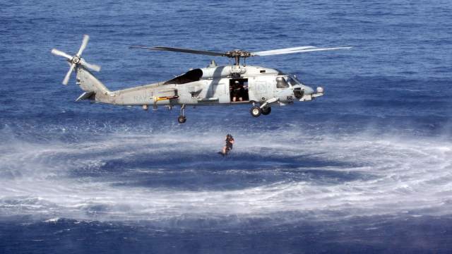 A helicopter hovers over the ocean as a person is lowered into the water, creating a circular ripple effect around them.