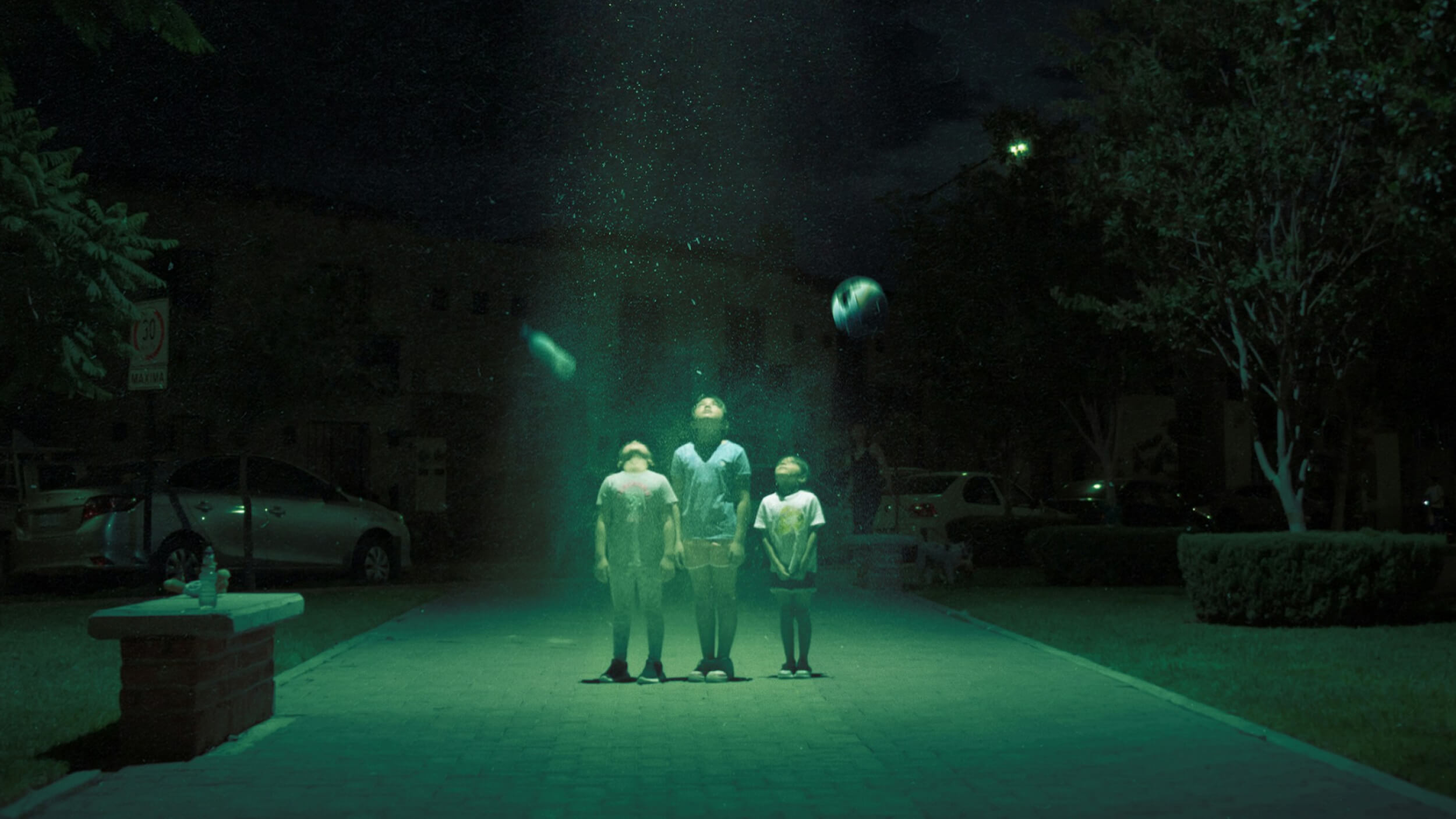 Three children stand under a mysterious green light in a suburban street at night, looking upward. Floating objects are illuminated around them.