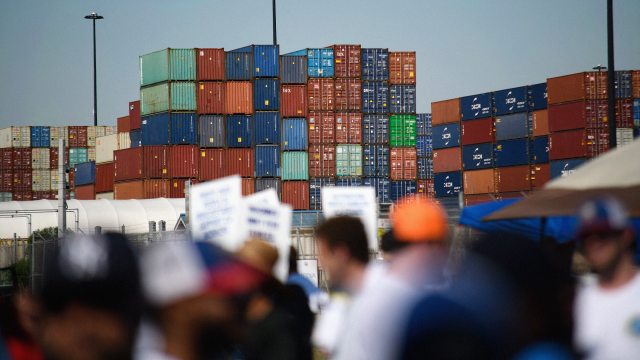 Stacks of colorful shipping containers are seen in the background, with out-of-focus people holding signs in the foreground.