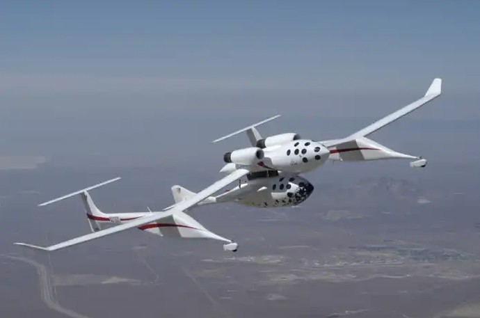 A white aircraft with a unique dual-fuselage design flying in a clear blue sky over a desert landscape.