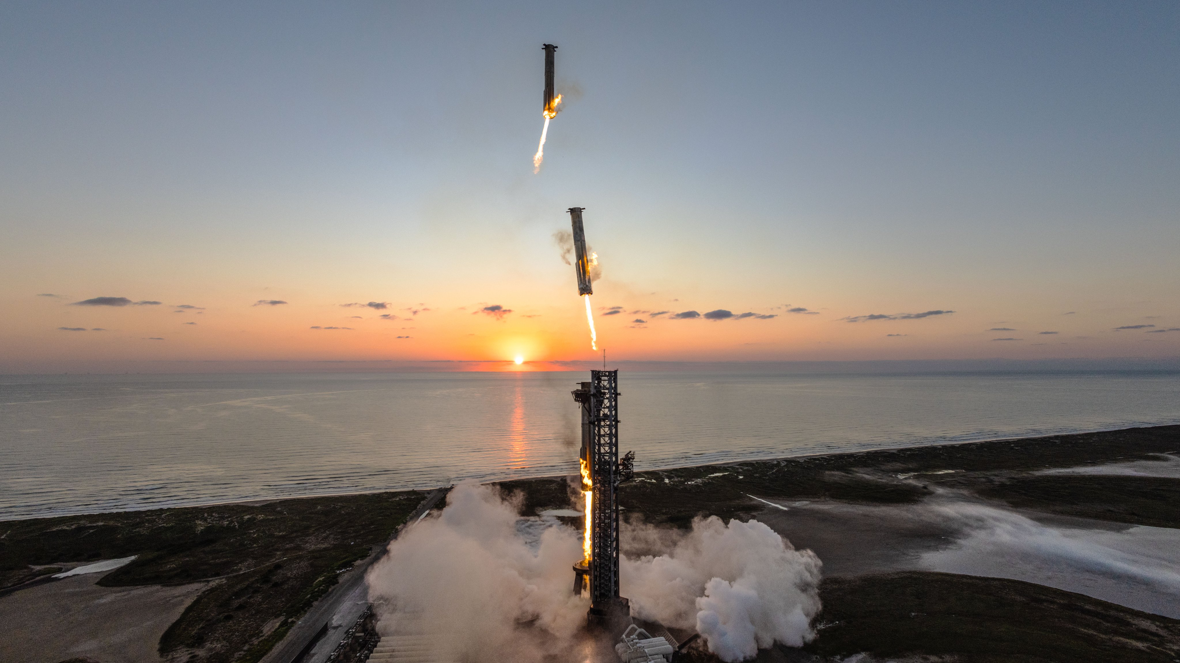 A rocket launching at sunrise, leaving a trail of smoke over a coastal landscape, with two boosters detaching and descending.