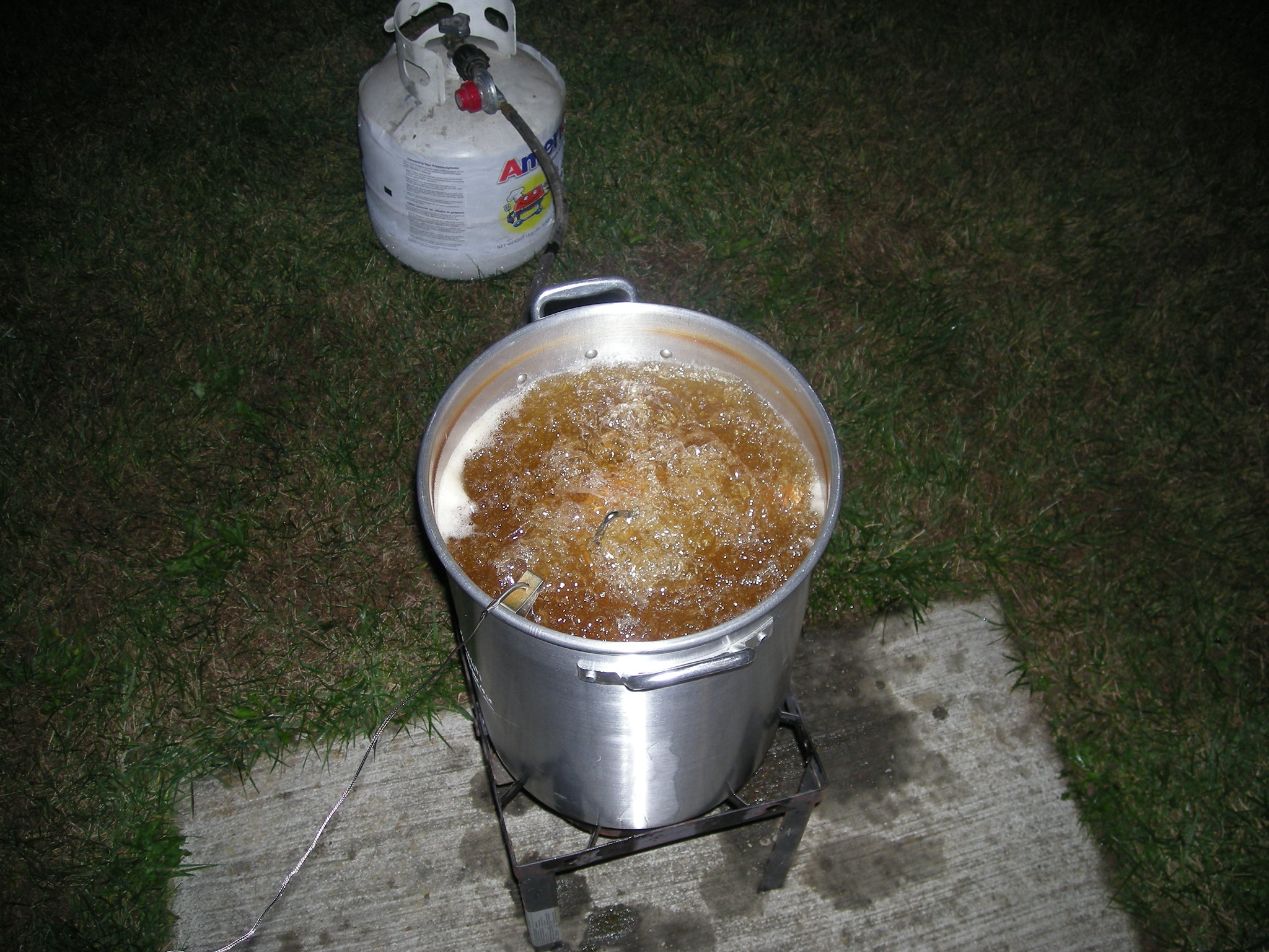 A large pot of oil deep frying a frozen turkey outdoors on a propane burner, with a propane tank nearby resting on grass and concrete.