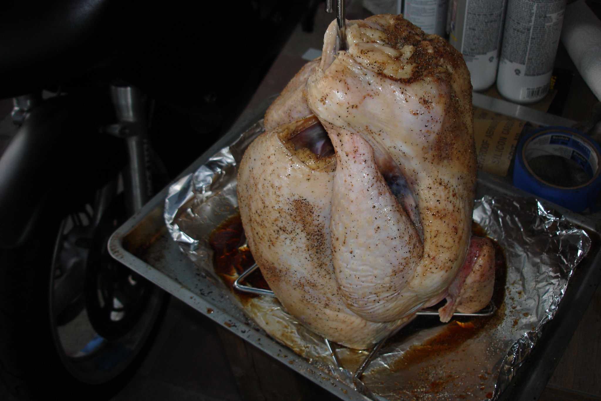 A seasoned whole chicken stands upright on a metal stand in a baking pan lined with foil, reminiscent of preparing to deep fry a frozen turkey.