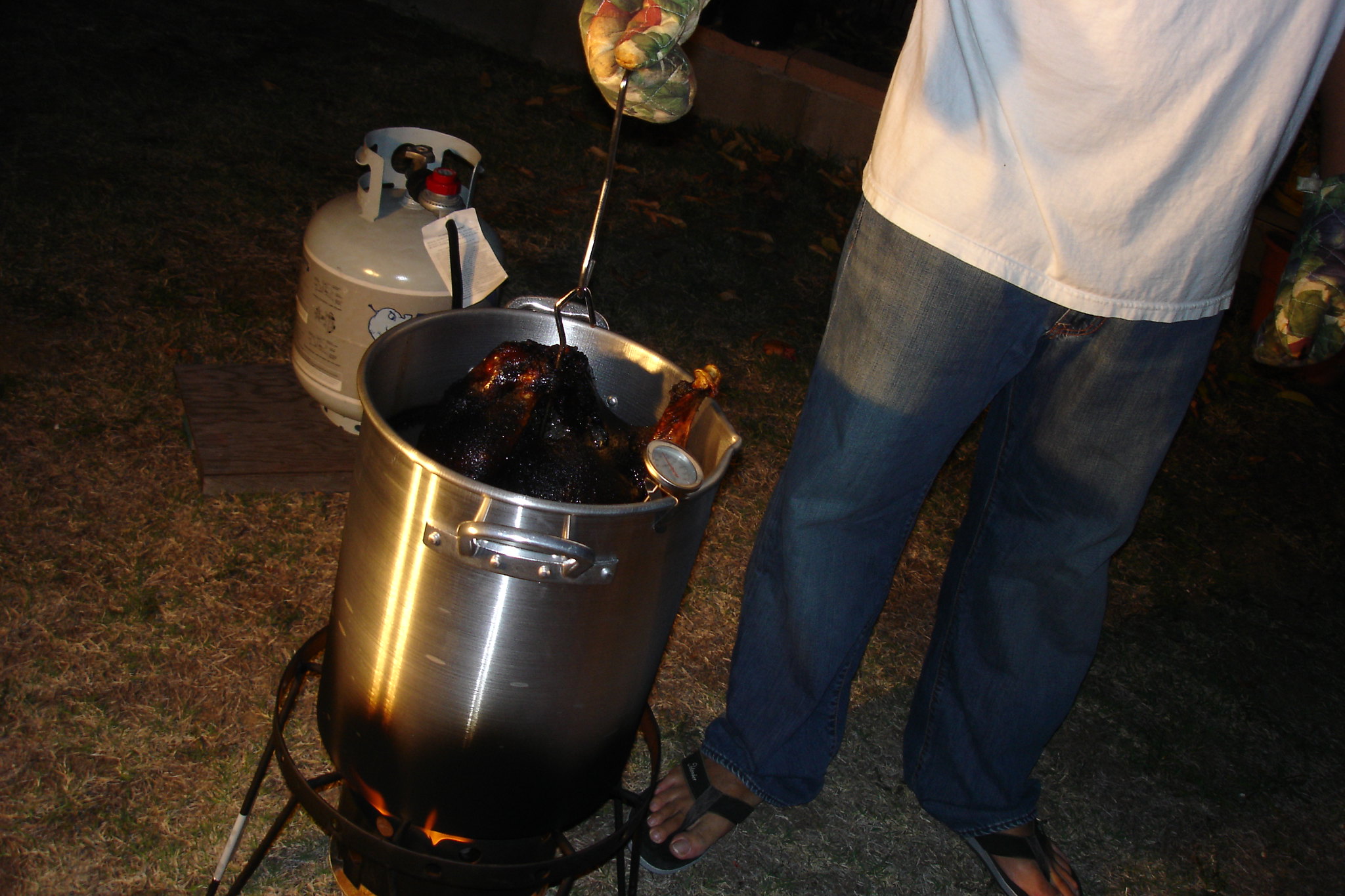 A person cautiously uses metal tongs to deep fry a frozen turkey in a large outdoor pot on a gas burner.