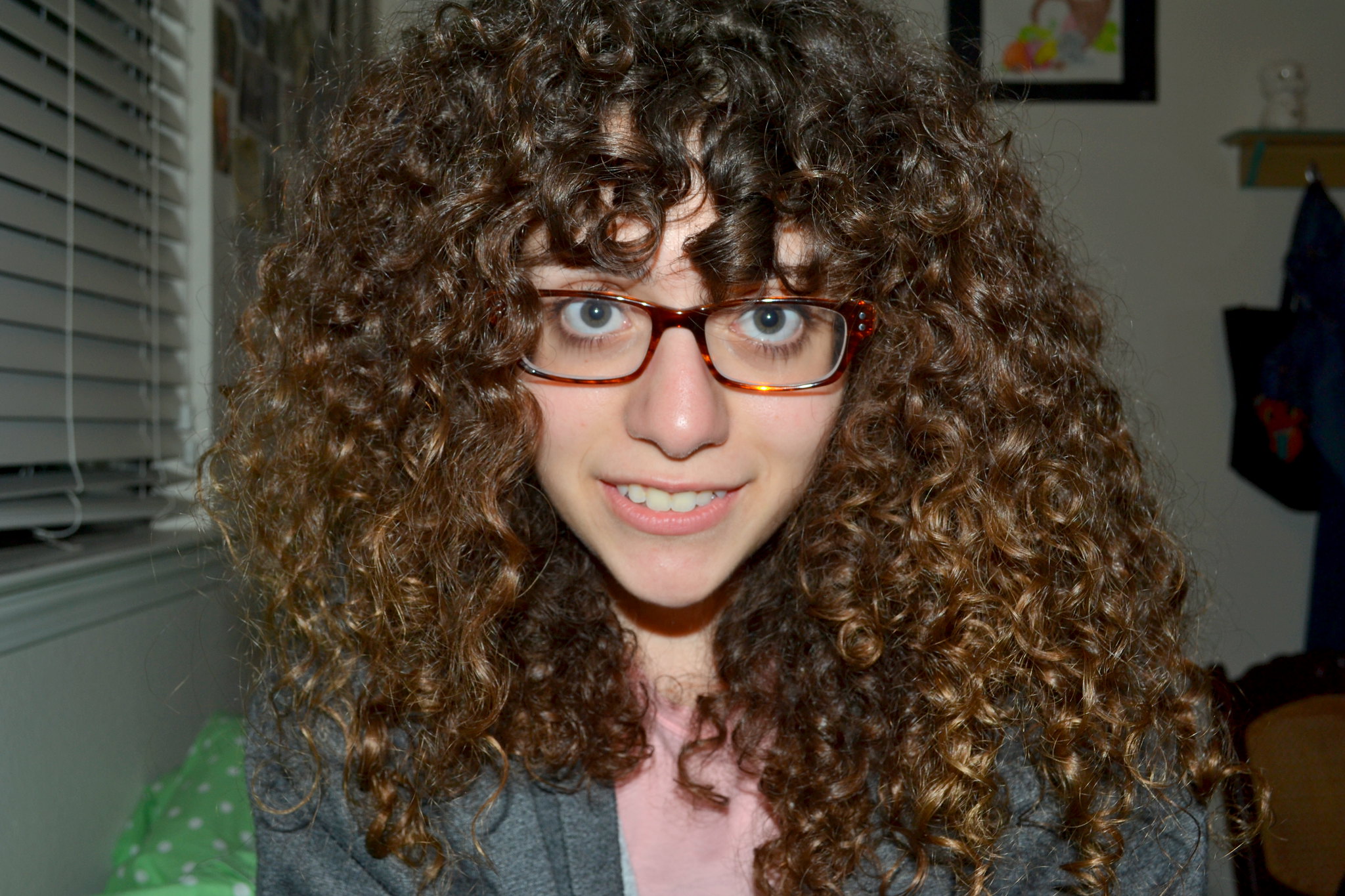 A person with curly hair and glasses smiles at the camera in a room with a window and wall decorations.