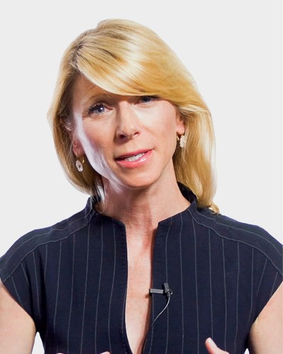 Person with blonde hair, wearing a pinstripe top and a clip-on microphone, speaking in front of a white background.